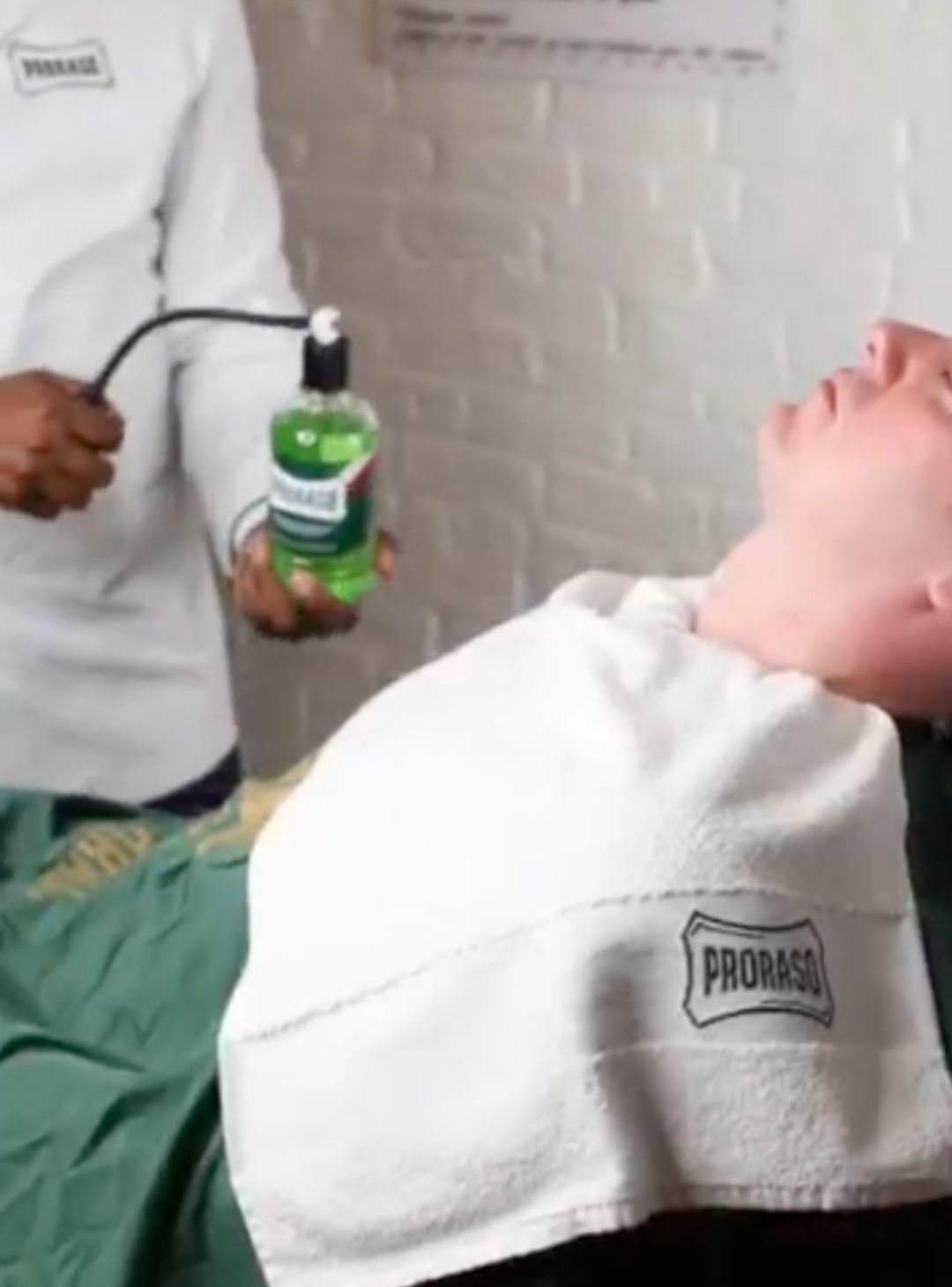 Client sitting in barber chair with Proraso towel over his him and a cape with a barber spraying After Shave Lotion