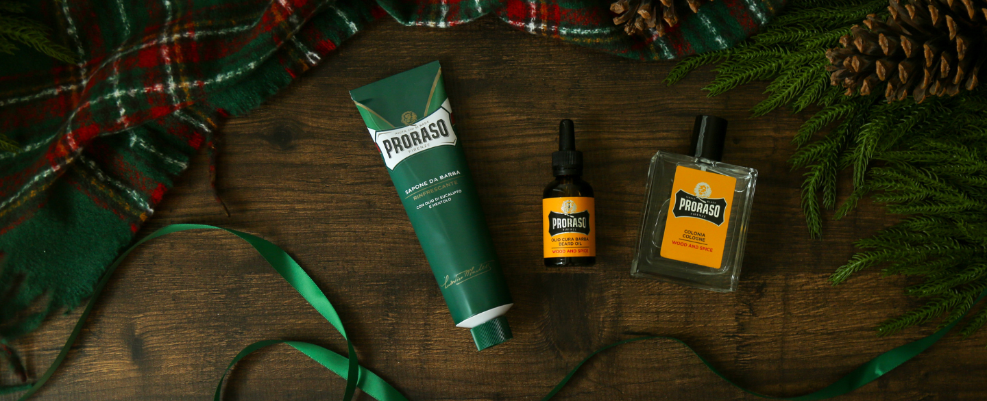 Tube of Refreshing Shave Cream, Wood & Spice Beard Oil and Cologne on a wooden floor with a plaid blanket and greenery in the background