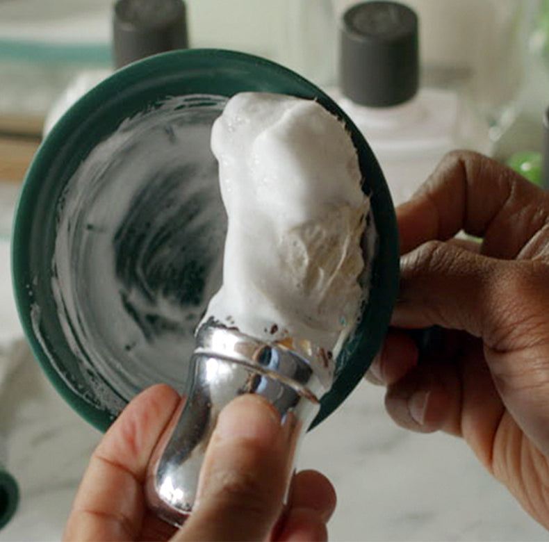 Man making thick lather with Professional Shave Brush in a green shave mug with two bottles of After Shave Balm in background
