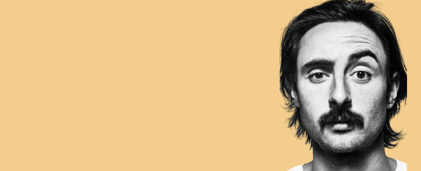 Man with messy short hair and a moustache in front of a yellow background