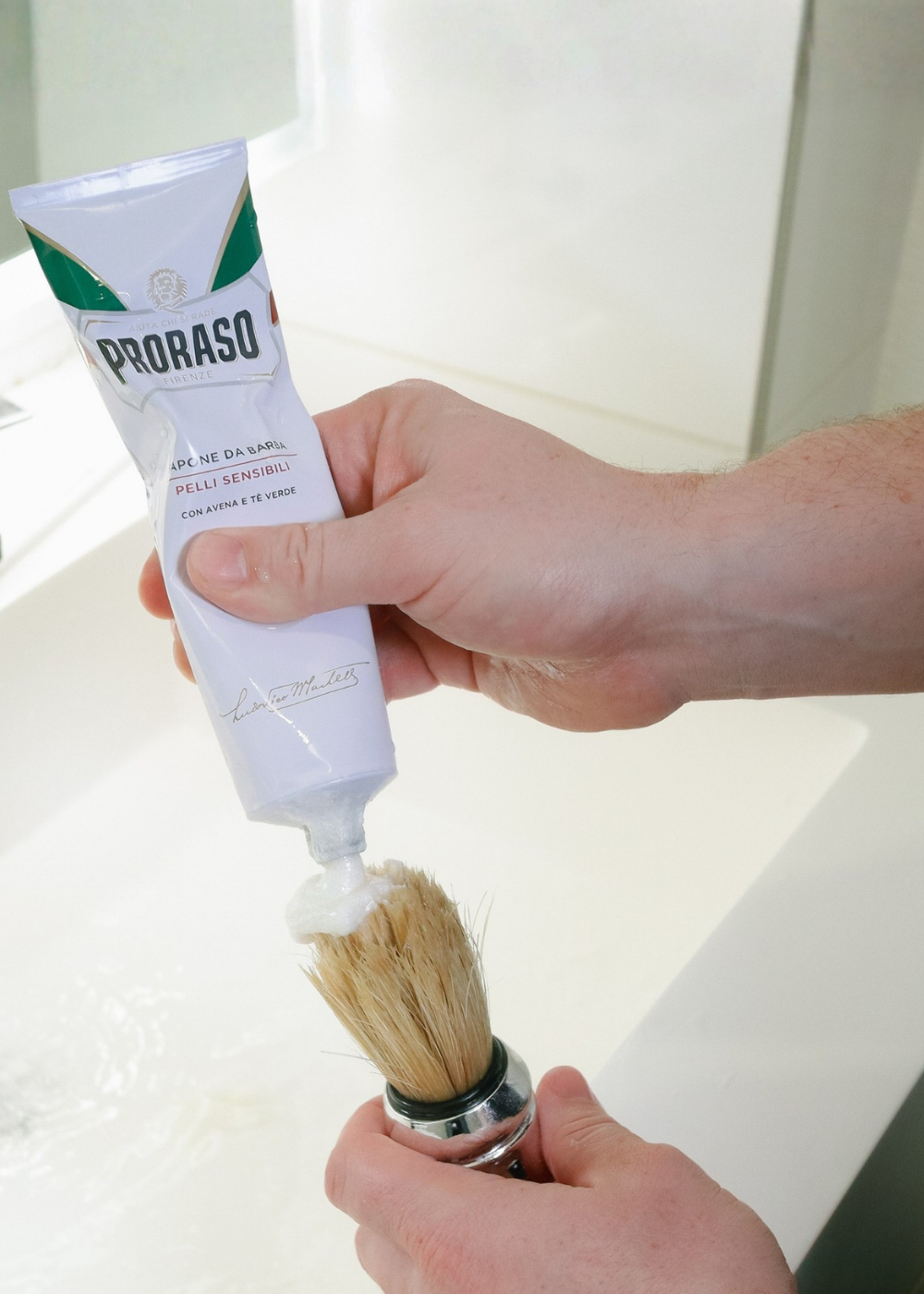 Man squeezing Sensitive Shave Cream Tube on our Professional Shave Brush in a white bathroom.