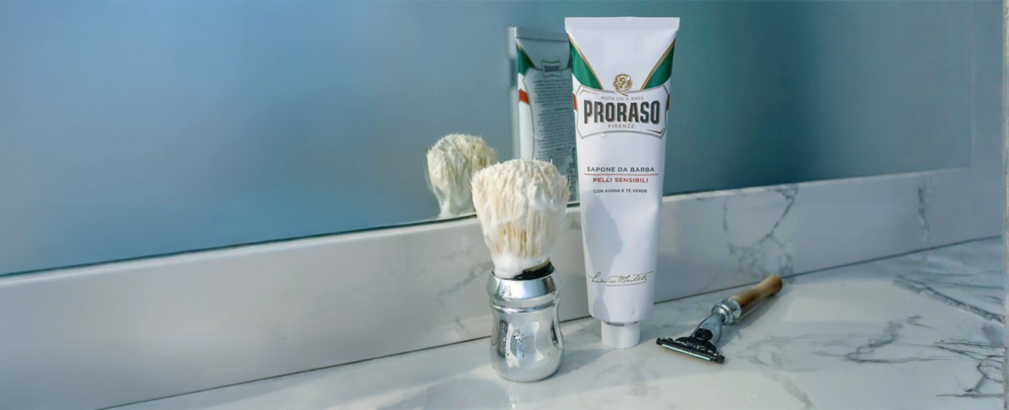 Tube of Sensitive Shave Cream with lathered Professional Shave Brush and horn razor on a white marble bathroom counter.