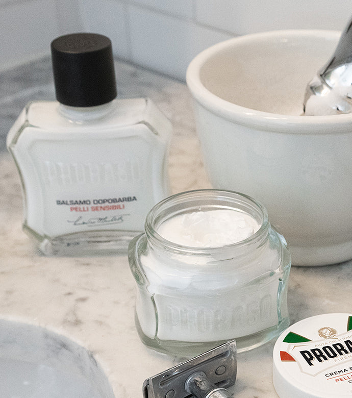 Proraso Sensitive After Shave Balm, Proraso Sensitive Pre-Shave Cream jar open, shave bowl with Proraso Professional Shaving Brush inside and safety razor sitting on bathroom counter.