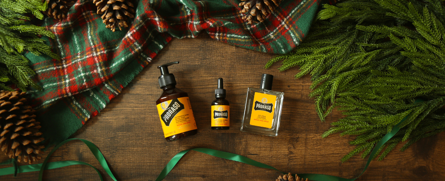 Wood & Spice Beard Wash, Beard Oil and Cologne on a wooden floor surrounded by a plaid blanket, green ribbon and tree foliage 