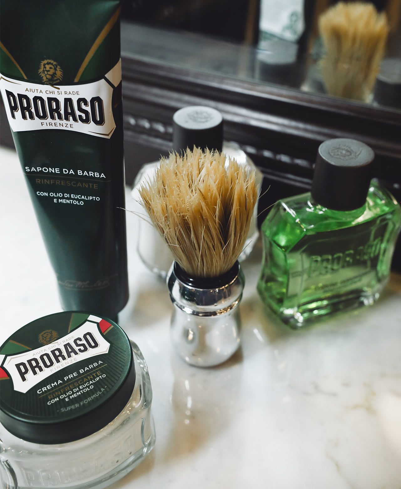 From left to right Refreshing line products sitting on a white marble barbershop counter Pre-Shave Cream, Shave Cream Tube, Professional Shave Brush, After Shave Balm and After Shave Lotion