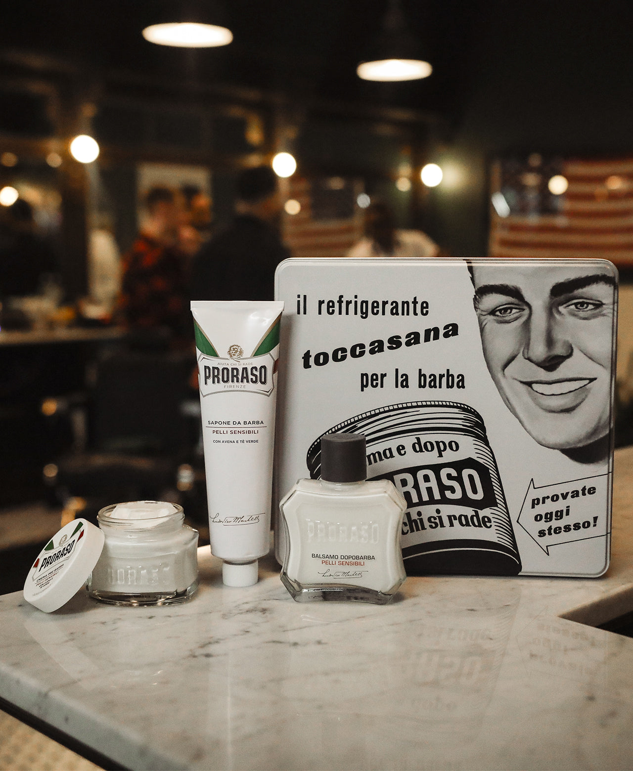 Vintage Toccassana Tin sitting on white marble counter with contents of set: Pre-Shave Cream, Shave Cream Tube and After Shave Balm with barbershop scene in background 