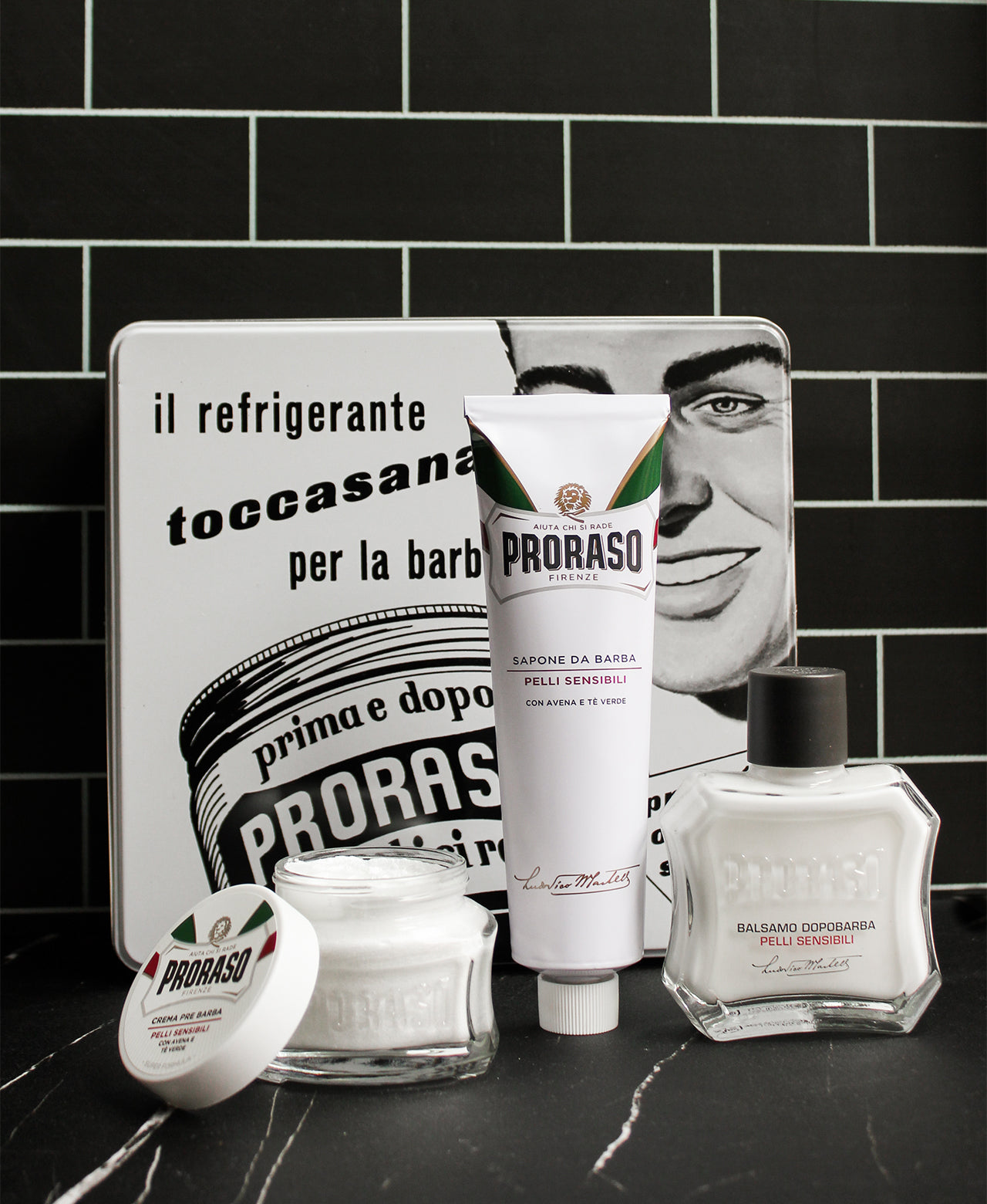 Contents of Toccassana Vintage tin in front of box: Pre-Shave Cream with jar open, Shave Cream Tube and After Shave Balm sitting on a black marble table in front of a black subway tile wall