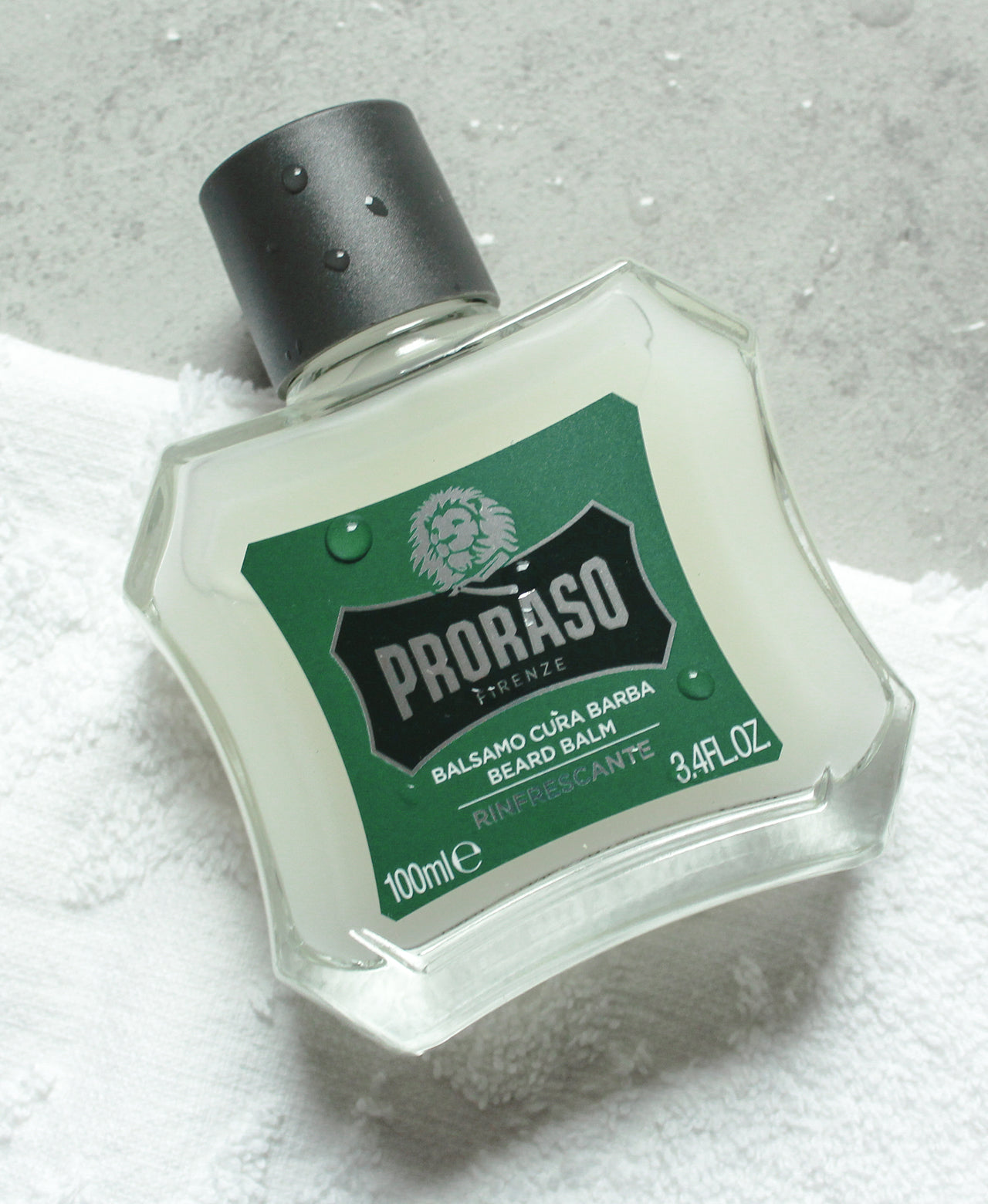 Bottle of Refresh Beard Balm with water drops sitting on a grey table and white towel