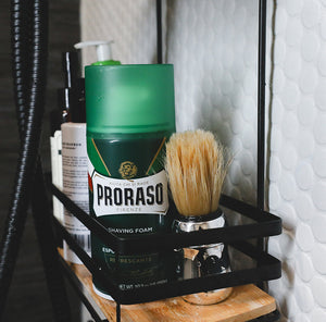 Refreshing Shave Foam can and Professional Shave Brush sitting in a shower on a caddy with next to a white tile wall