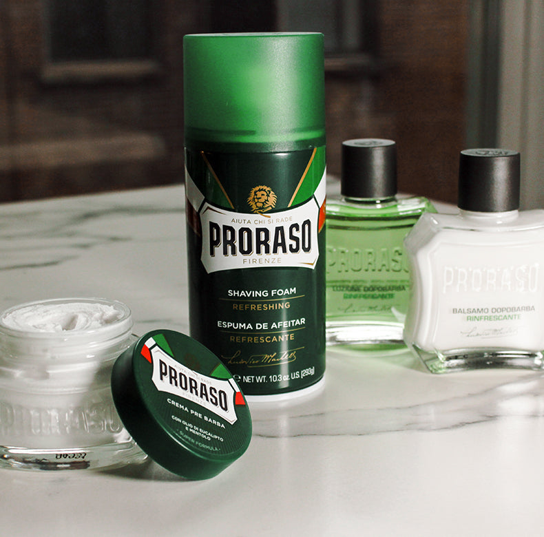 Refreshing formula lineup sitting on a white marble table: Pre-Shave Cream jar with lid open to the side, Shave Foam, After Shave Lotion and After Shave Balm