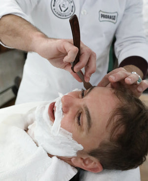 Man in barber chair with thick lather of Shave Cream Tube on his face, with a barber shaving his face in the background