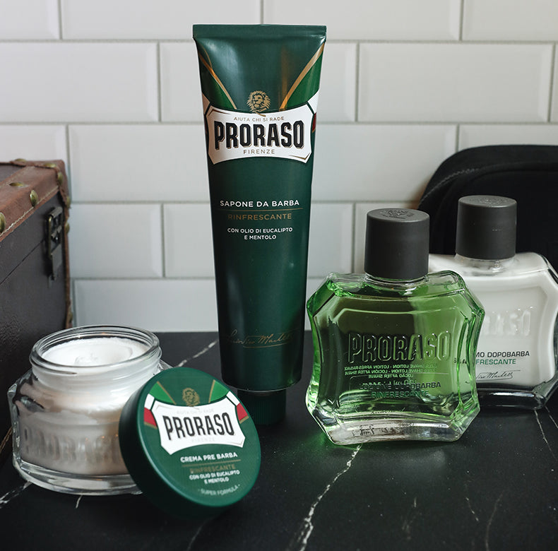 Refreshing formula lineup from left to right: Pre-Shave Cream jar open with lid facing front, Shave Cream Tube, After Shave Lotion and After Shave balm sitting on a black marble table with a suitcase and dopp kit in front of a white subway tile wall.