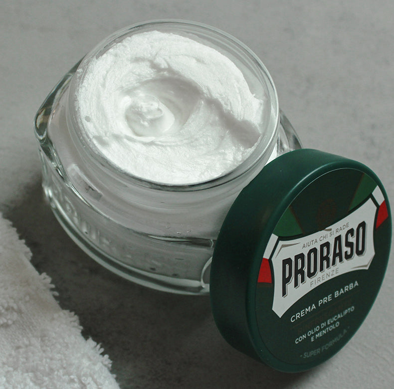 Refreshing Pre-Shave Cream jar open and lid tilted on right side of jar, sitting on a grey table with white towel in corner.