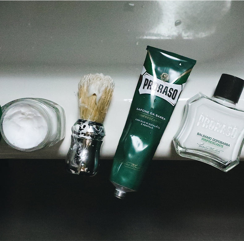 Refreshing formula three steps laying on side of bathtub from left to right, open jar of Pre-Shave Cream, Professional Shave Brush with lather on it, Shave Cream Tube and After Shave Balm 