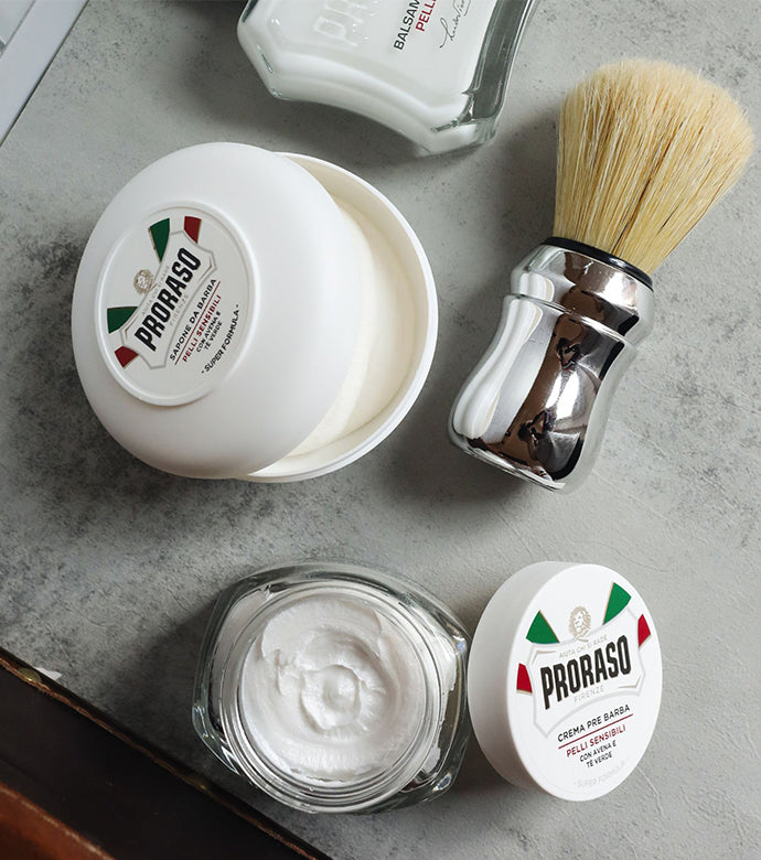 Sensitive formula products sitting on grey table: Pre-Shave Cream with jar open and lid sitting on side of jar, Shave Soap in a Bowl with lid partially off jar, of of a After Shave Lotion bottle in view and Professional Shave Brush to the right