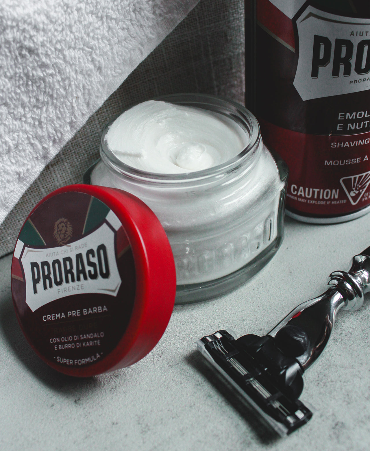 Jar of Coarse Beard Pre-Shave Cream with lid open to show the thick texture sitting next to a Coarse Shave Foam on a grey table with a Mach 3 razor in the front.