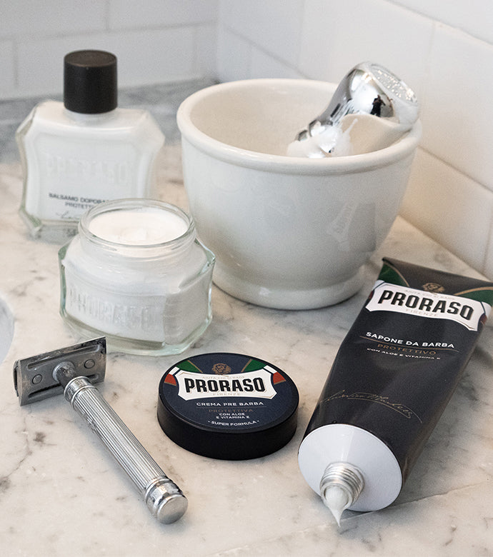 Protective formula lineup on white marble bathroom counter lineup. From front to back: open Shave Cream Tube with lid off, silver double edge safety razor, open jar of Pre-Shave Cream with lid in front, white shave jar with a Professional Shave Brush in bowl, and After Shave Balm with a white tile background