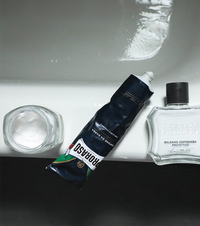 Protective formula lineup on the edge of a bathtub from left to right open jar of Pre-Shave Cream, Shave Cream Tube without lid and After Shave Balm