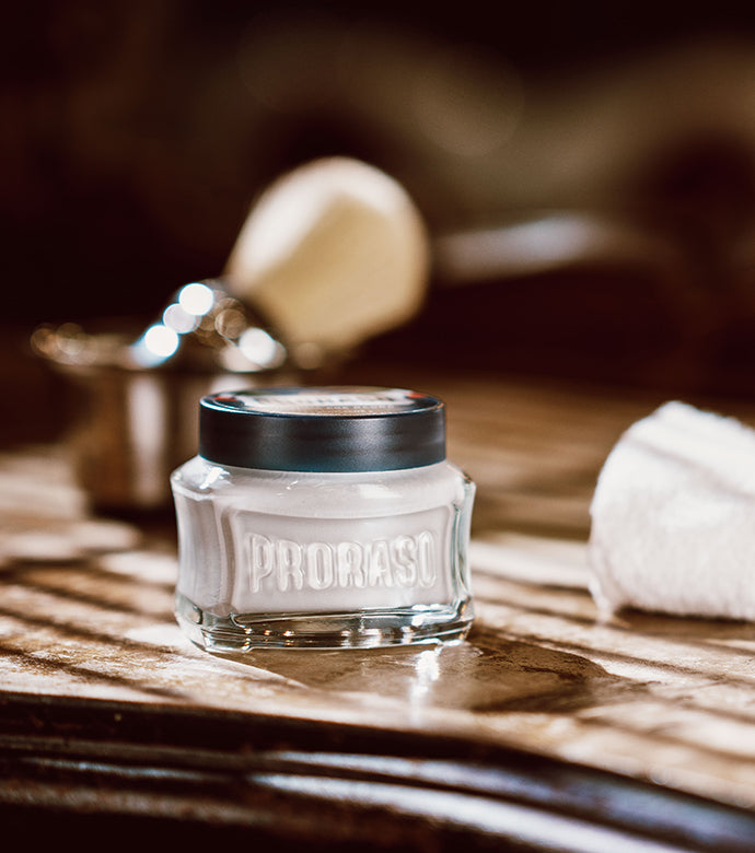 Protective Pre-Shave Cream jar on dark wood table with Professional Shave Brush in silver bowl and white towel in background 