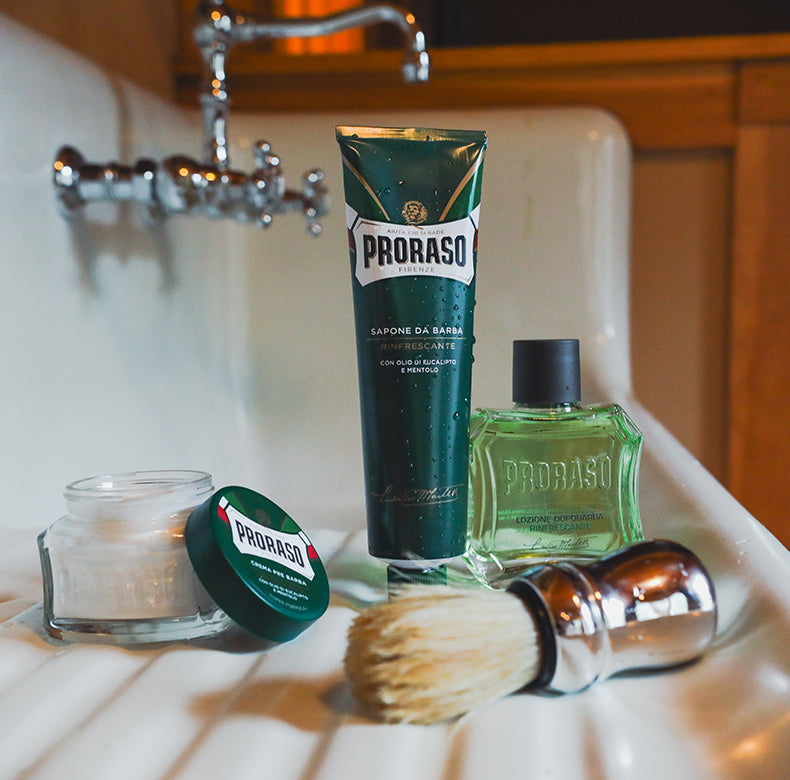 Refreshing formula lineup sitting on a vintage sink counter from left to right Pre-Shave Cream Jar open with lid sitting on side of jar, Shave Cream Tube and After Shave Lotion with a Professional Shave Brush sitting in the front.