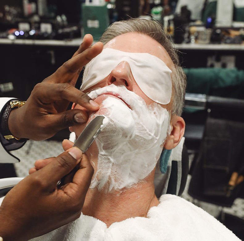 Refresh Shave Cream Tube applied to a man's face sitting in a barber chair while a barber uses a safety razor to shave his face