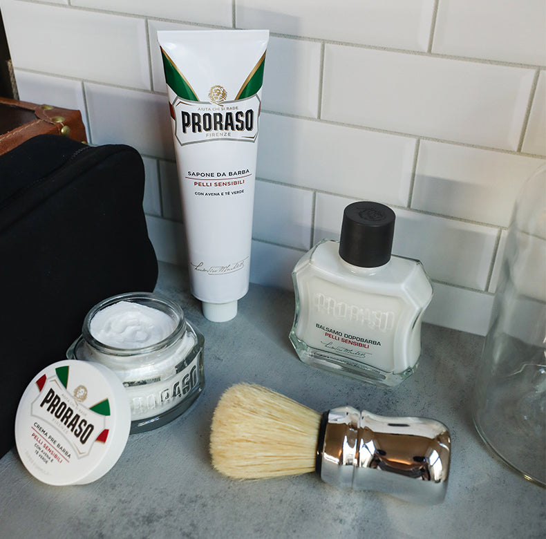 Sensitive Formula lineup from left to right: Pre-Shave Cream with jar open to see thick texture and lid balanced on the site, Shave Cream Tube, After Shave Lotion and Professional Shave Brush in front sitting on a grey table with white subway tile wall