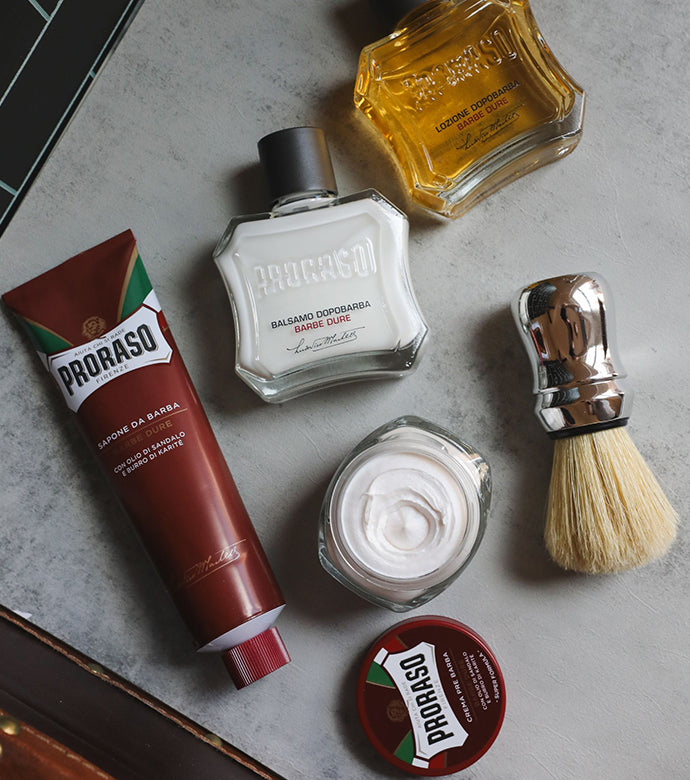 Coarse Beard formula lineup sitting on a grey table products left to right Shave Cream Tube, open jar of Pre-Shave Cream with lid in front, After Shave Balm and After Shave Lotion with Professional Shave Brush sitting in front of a black tile wall