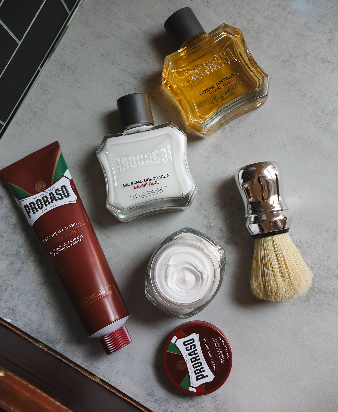 Coarse Beard formula lineup spread out on a grey table from left to right: Shave Cream Tube, Pre-Shave Cream with lid open to show texture, After Shave Balm, Professional Shave Brush and After Shave Lotion with a black subway tile wall 