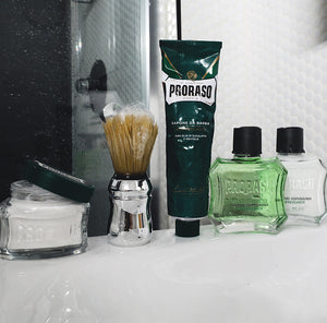 Refreshing formula lineup sitting on edge of bathroom sink from left to right Pre-Shave Cream, Professional Shave Brush with lather suds, Shave Cream Tube, After Shave Lotion and After Shave Balm with white wall and shower in the background