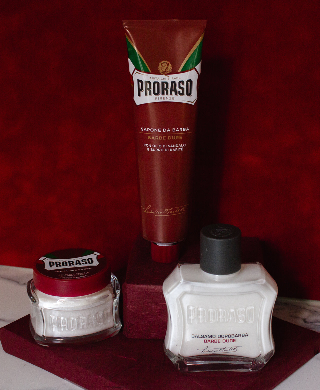 Nourishing formula lineup on graduated red steps from left to right: Pre-Shave Cream, Shave Cream Tube and After Shave Balm in front of a red background 