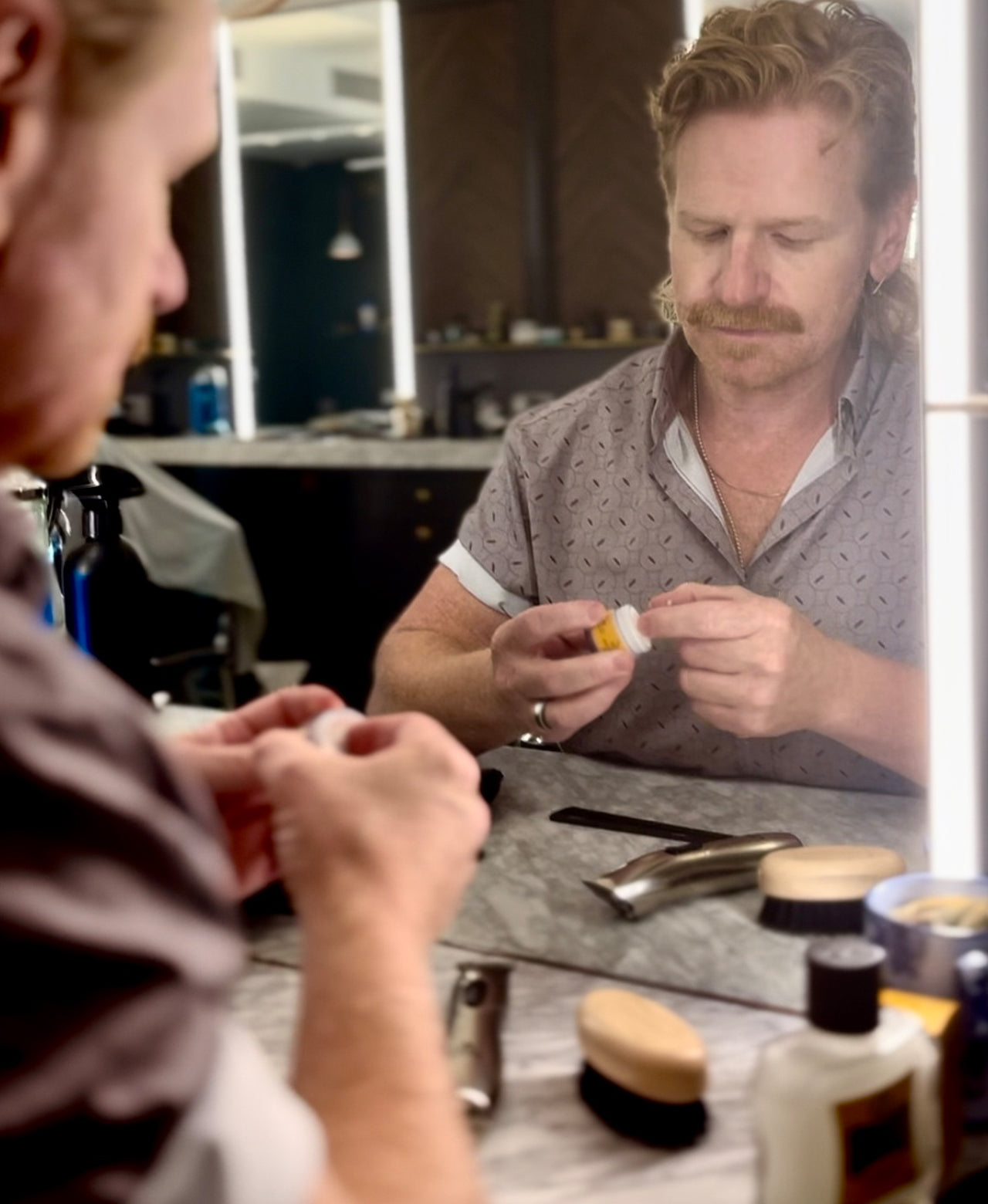 Man scooping out a small amount of Moustache Wax onto his finger in front of a barber mirror with various styling tools like trimmers, comb, Moustache and Beard Brush and Beard Balm on the counter.