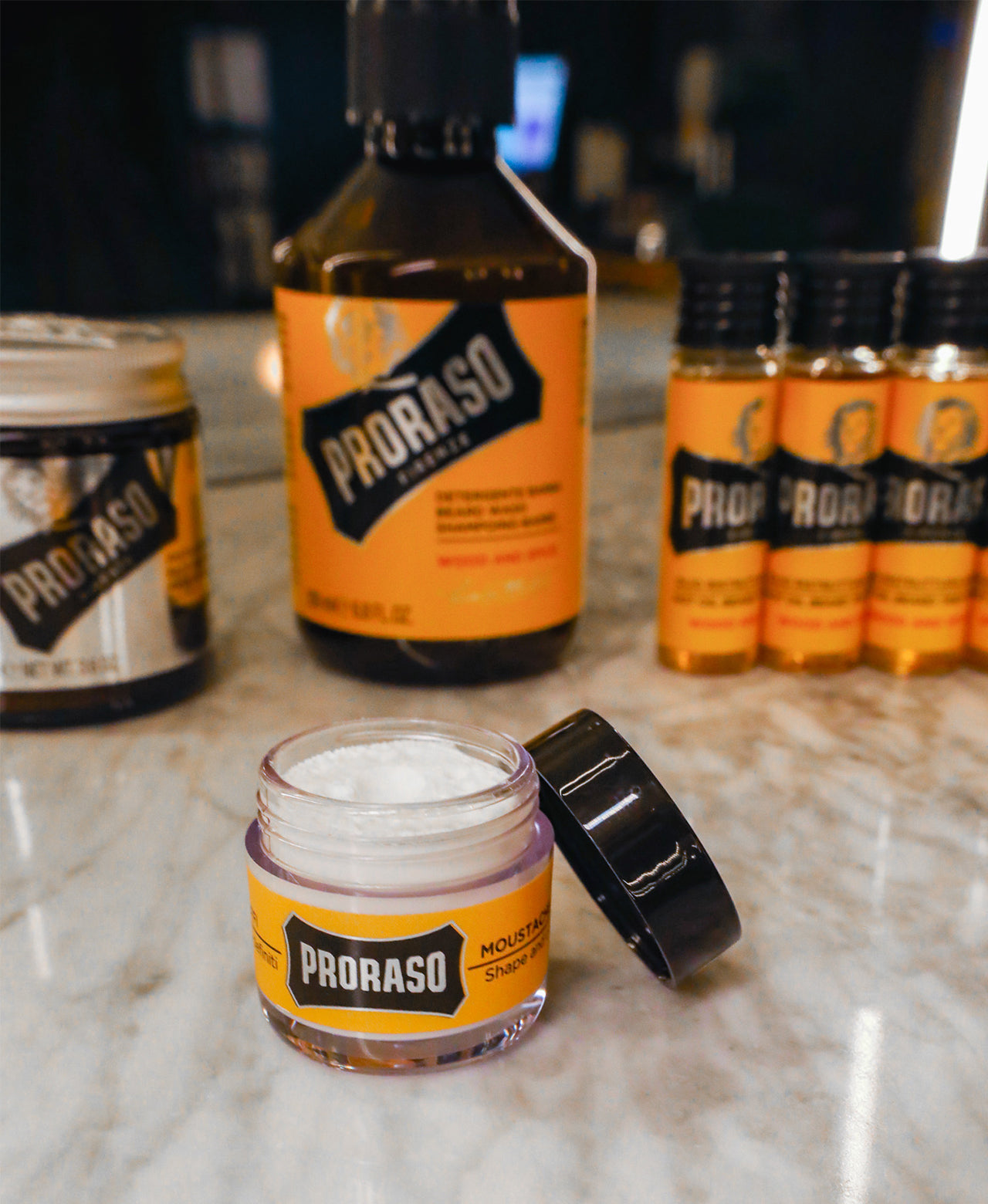 Arrangement of beard product: Moustache Wax in front with jar open and lid to the side. In the back row left to right: Exfoliating Beard Paste & Facial Scrub, Wood & Spice Beard Wash and three vials of Hot Oil Beard Treatment on a white marble counter with mirror in the background 