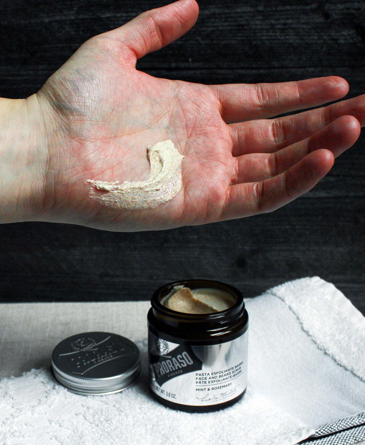 Palm of hand with swipe of Exfoliating Beard Paste and Facial Scrub to show thick texture with towel on grey table in front of a black wooden wall