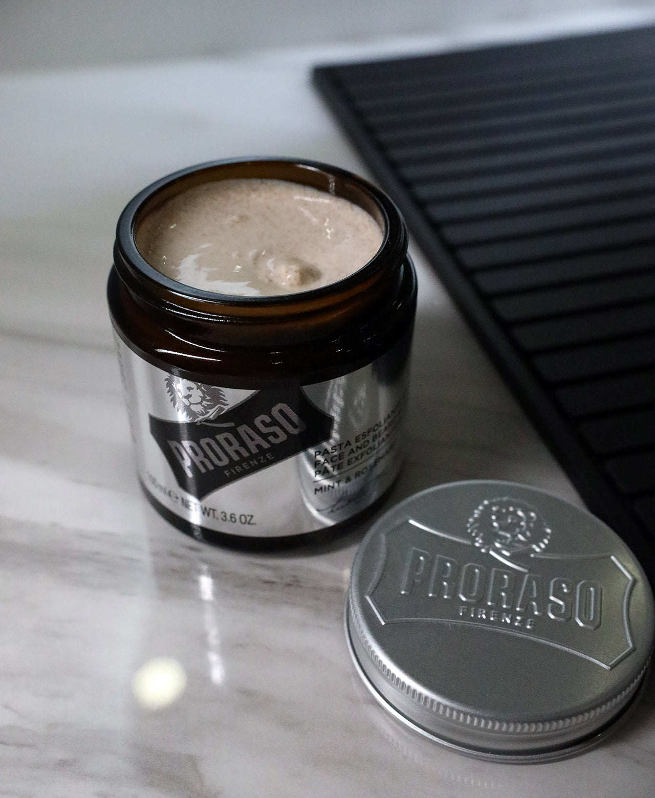 Jar of Exfoliating Beard Paste and Facial Scrub with lid open in front on a white marble barbershop counter