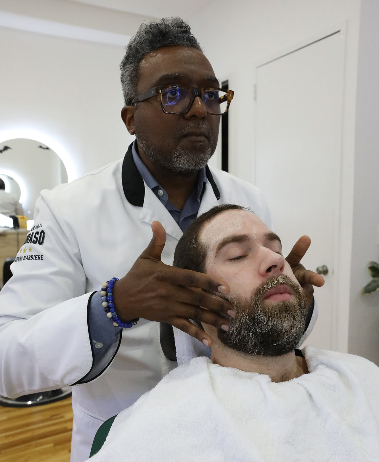 Barber applying Exfoliating Beard Paste and Facial Scrub to client in a barber chair with barbershop in background