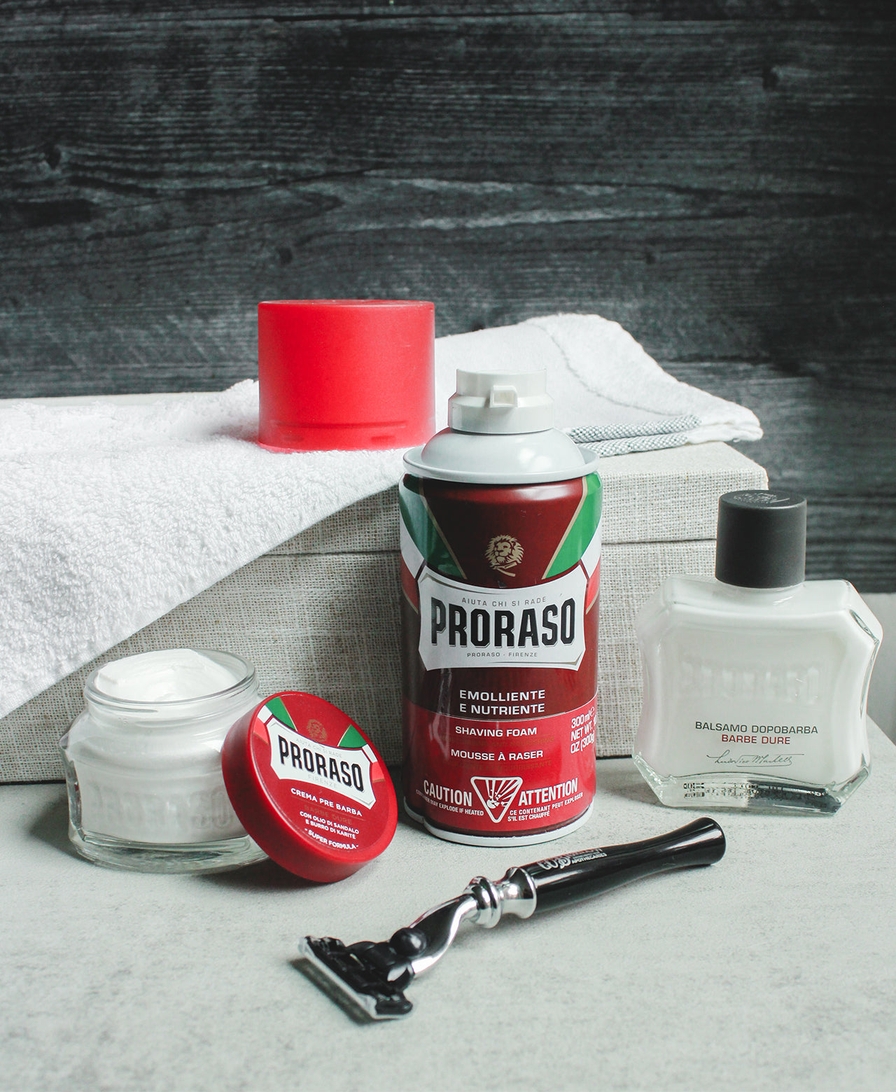 Nourishing formula lineup from left to right: Pre-Shave Cream with lid off, Shave Foam, After Shave Balm with a mach 3 razor in the front.The background shows a box with a white towel partially covering it with a dark wood backdrop.