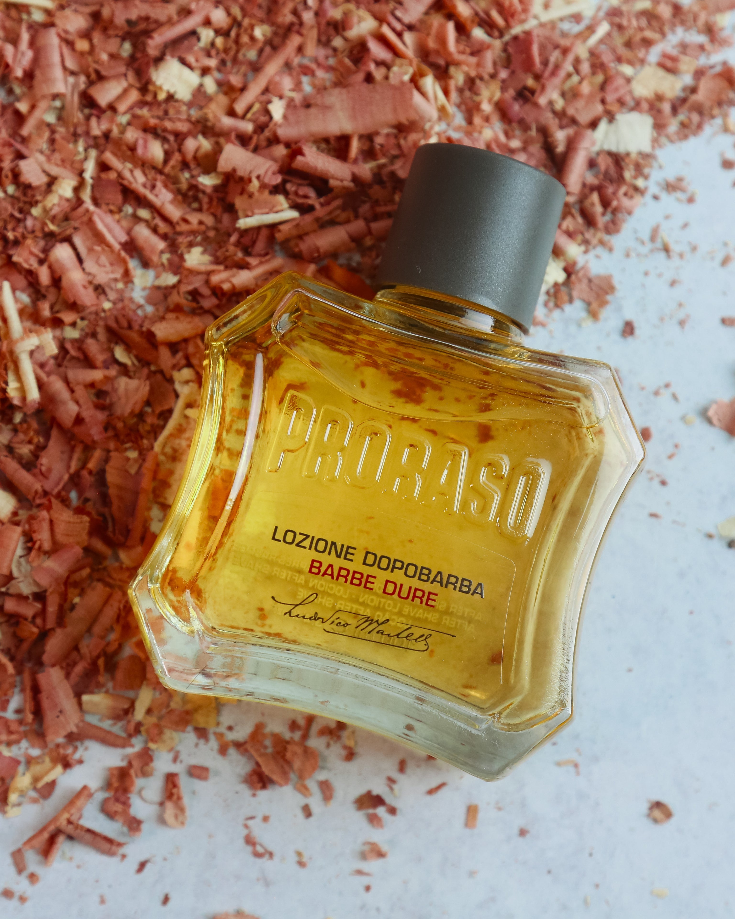 A bottle of Proraso Coarse Beard After Shave Lotion, filled with amber-colored liquid and featuring a black cap, styled on a white surface surrounded by sandalwood shavings.