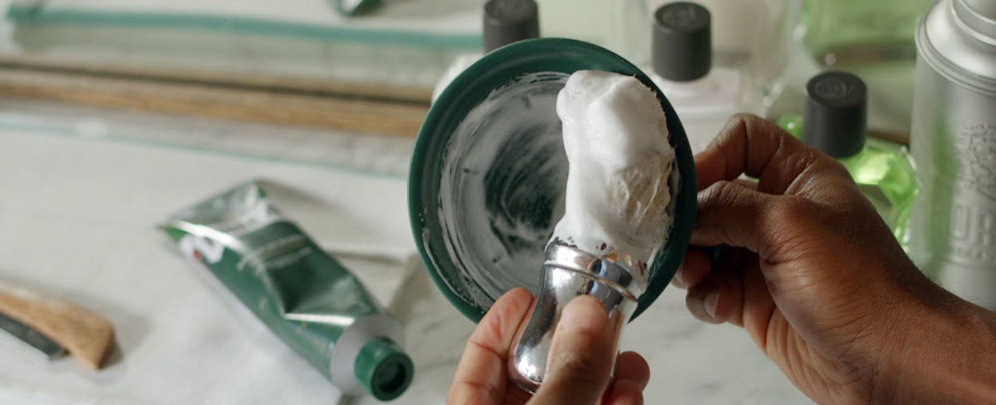 Barber hands building a thick lather in a green Proraso shave mug. The background shows Proraso Refreshing Formula Shave Cream Tube, After Shave Balm, After Shave Lotion and shaker