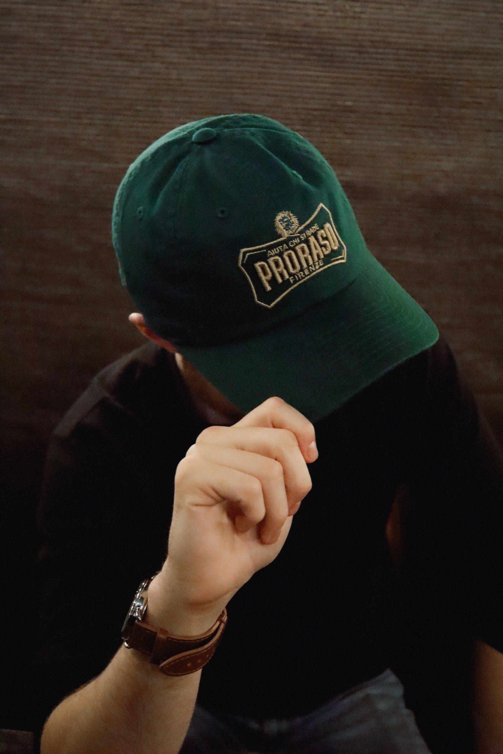 Man wearing green Proraso cap with gold logo tilted to the side with his hand on the brim