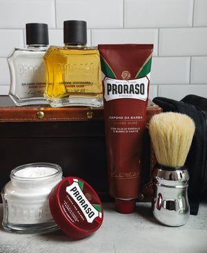 Nourishing Shave formula lineup: Pre-Shave Cream with open lid, Shave Cream Tube and Professional Shaving Brush in front. After Shave Balm and After Shave Lotion sit in the background on top of a vintage suitcase with a black dopp kit all in front of a white subway tile background 