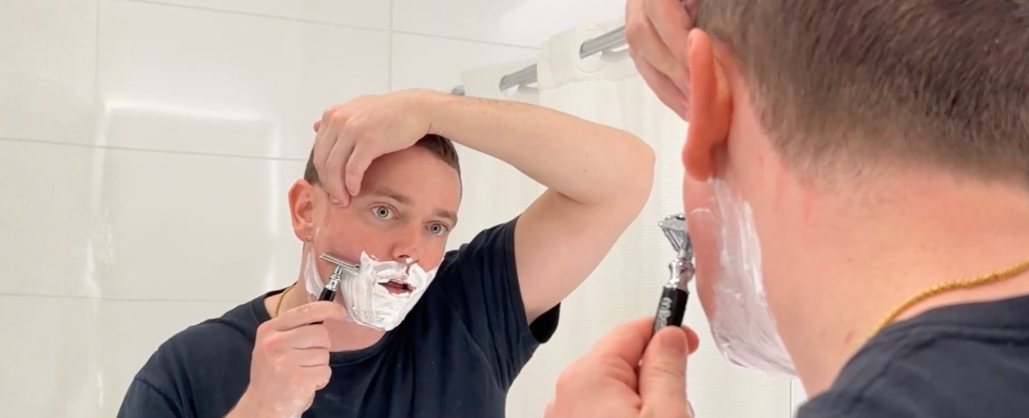 Man with brown hair shaving in front of a mirror in a white tile bathroom