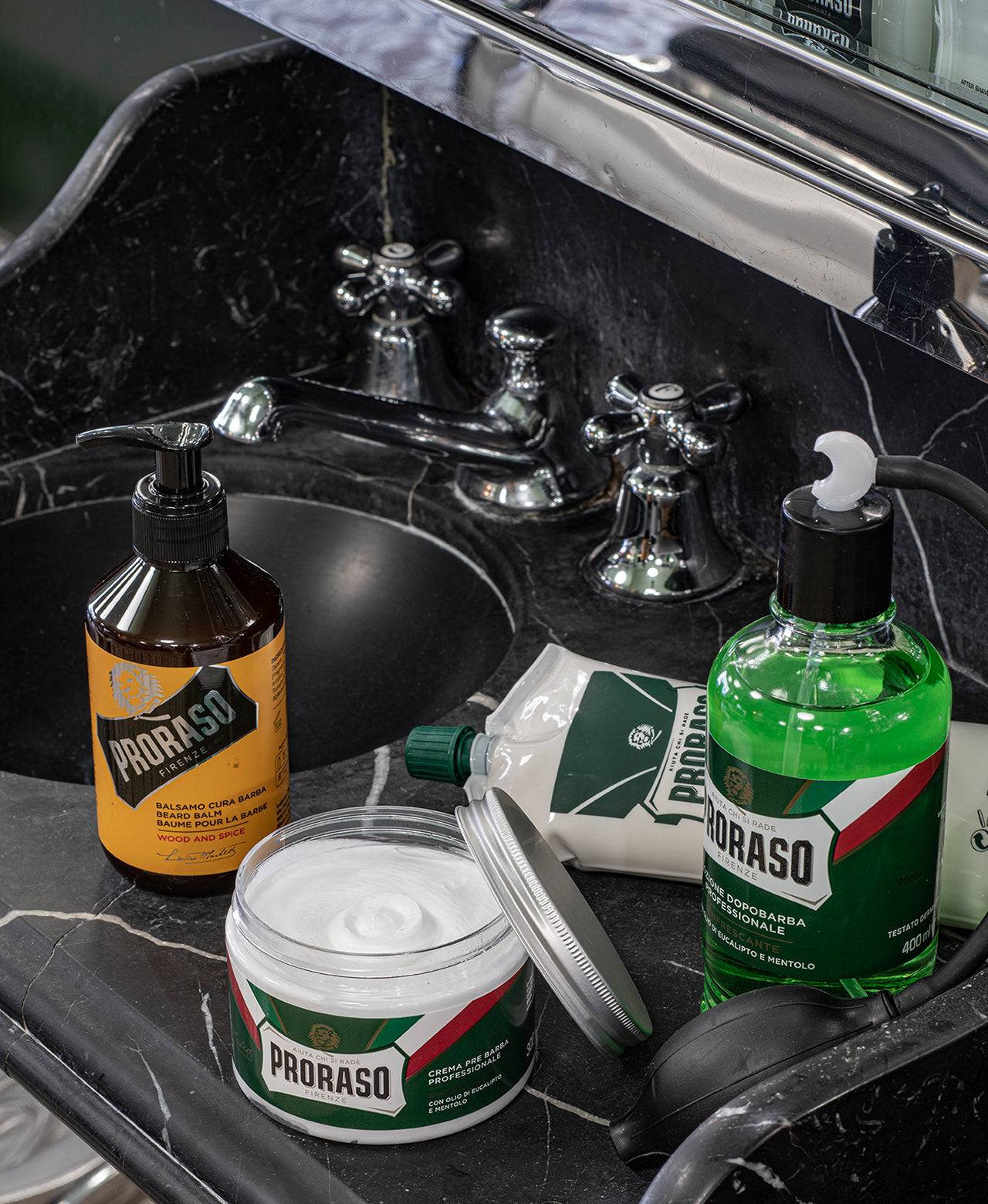 Professional size products sitting on a black marble sink counter from left to right Wood & Spice Beard Balm, open jar of Refresh Pre-Shave Cream, Refreshing Shave Cream Tube and After Shave Lotion with bulb