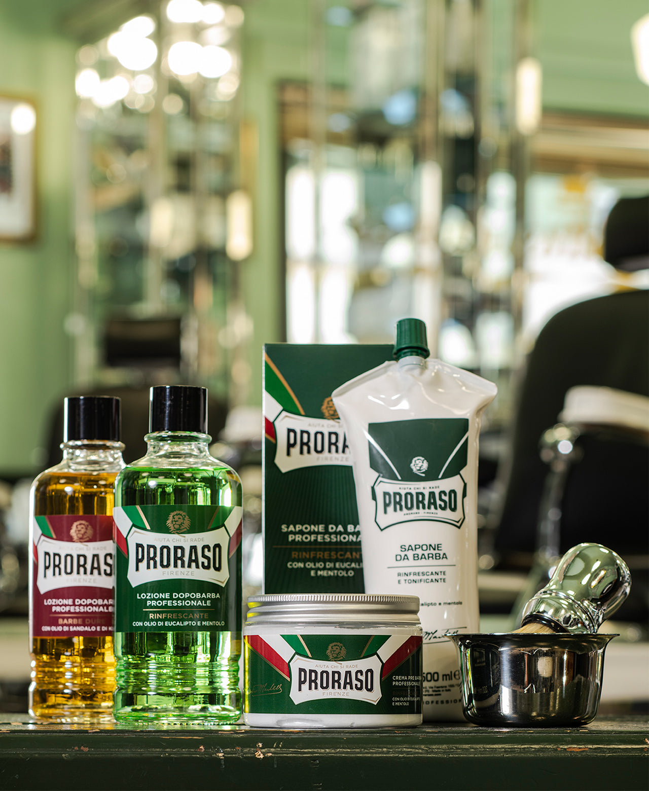 Professional size lineup from left to right Coarse Beard After Shave Lotion, Refresh After Shave Lotion, Pre-Shave Cream, Shave Cream with box and Professional Shave Brush in a shaving bowl. The background shows a Proraso green barbershop