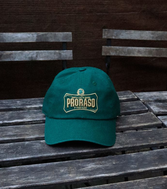 Green cap with Proraso logo in gold embroidered on the front sitting on a wooden table with chairs in the background