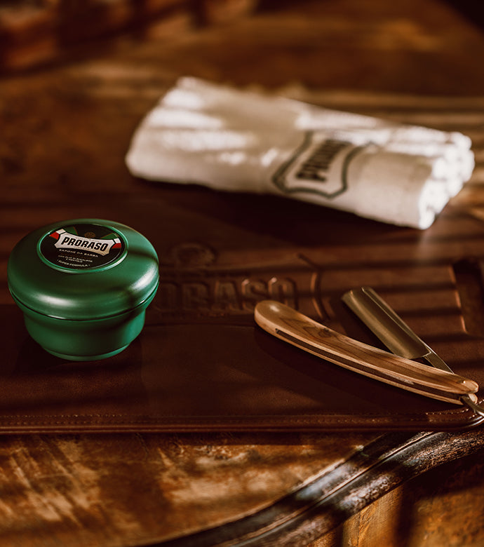 Refreshing Shaving Soap in a Bowl with lid closed sitting on a dark brown table with a straight razor and Proraso white towel 