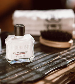 Bottle of Sensitive After Shave Balm with Moustache and Beard Brush sitting in the background and Proraso white towels on a brown wooden table