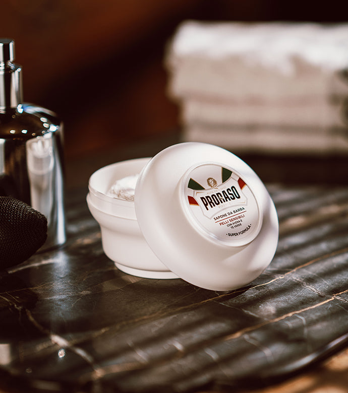 Sensitive Shaving Soap in a Bowl with open jar with barber tools in the background on dark brown wood table