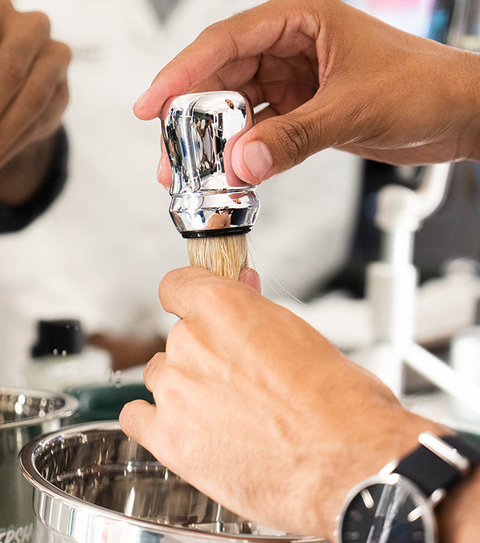 Hands squeeze excess water from a Proraso Professional Shaving Brush into a metal shave bowl.