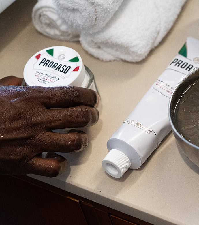 Proraso Sensitive line on bathroom counter, hand reaching to grab Proraso Sensitive Pre-Shave Cream with Proraso Sensitive Shaving Cream Tube next to shaving bowl.