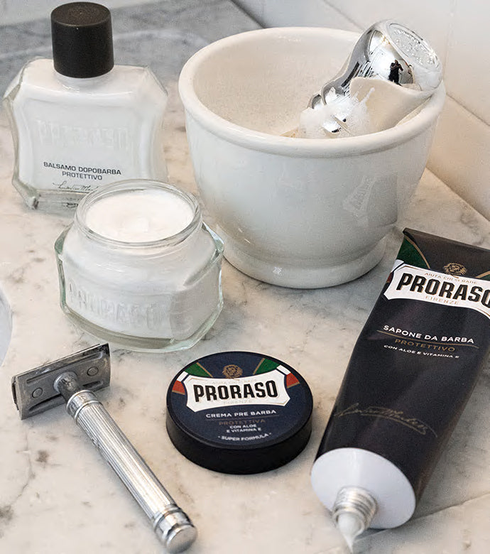 Proraso Protective Line on bathroom counter: Proraso Protective Aftershave Balm, Proraso Protective Pre-Shave Cream with lid off, Proraso Protective Shaving Cream Tube with cream squeezed out, with a safety razor, shave bowl and Proraso Professional Shaving Brush.